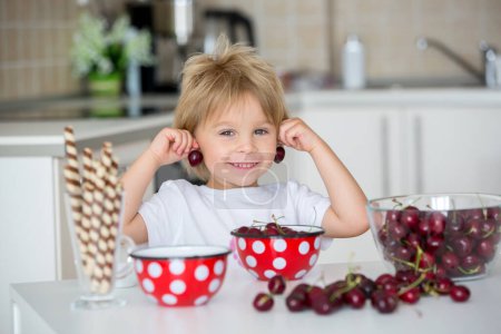Téléchargez les photos : Enfant blond mignon, tout-petit garçon, manger des cerises à la maison, s'amuser - en image libre de droit