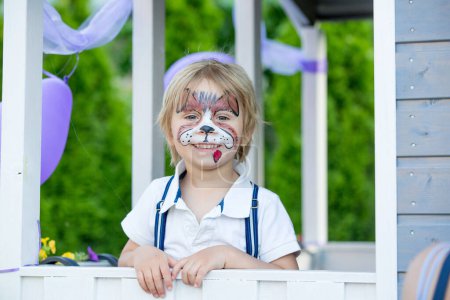 Foto de Little toddler baby boy, child with painted face as a dog, playing with pet dog in the garden on birthday party - Imagen libre de derechos