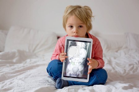 Photo for Toddler child, blond boy, holding x-ray picture on tablet of child body with swallowed magnet showing, child swallow dangerous object - Royalty Free Image