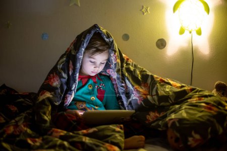 Téléchargez les photos : Cute preschool child, playing on tablet at home in bed in his pajamas - en image libre de droit