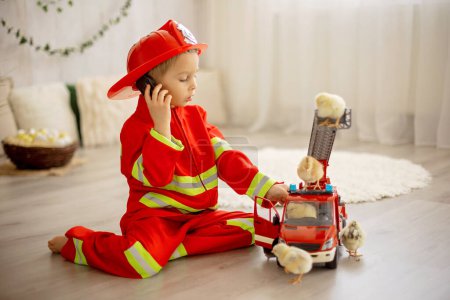 Téléchargez les photos : Petit enfant en bas âge, jouant avec le jouet de voiture de camion de pompier et les petites poussins à la maison, amis d'enfant et d'animal de compagnie jouant - en image libre de droit