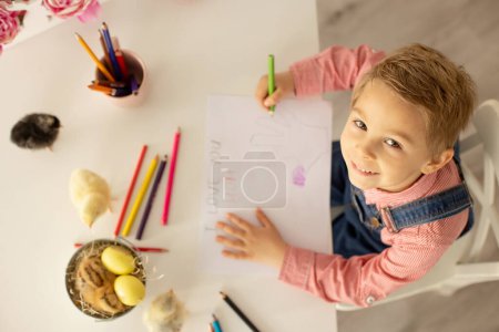 Foto de Cute boy, child in red shirt, drawing picture for Mothers day, little chicks on the table playing - Imagen libre de derechos