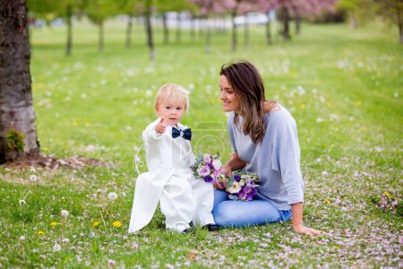 Téléchargez les photos : Beau tout-petit garçon, vêtu de smoking blanc, tenant un magnifique bouquet de fleurs pour la fête des mères, souriant joyeusement et le donnant à maman dans le parc - en image libre de droit