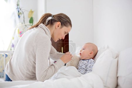 Foto de Lindo niño enfermo, bebé, quedarse en la cama, mamá dándole medicamentos y revisando si tiene fiebre - Imagen libre de derechos