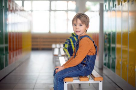 Foto de Dulce niño rubio de pie frente a un casillero en el jardín de infantes o el pasillo de la escuela, invierno - Imagen libre de derechos