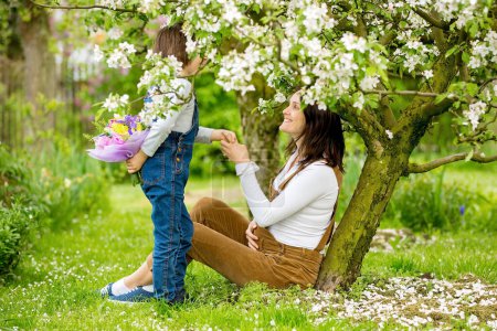 Foto de Mujer embarazada joven, recibiendo ramo de flores de colores de su hijo para el Día de las Madres, sentado en un hermoso jardín floreciente de primavera - Imagen libre de derechos