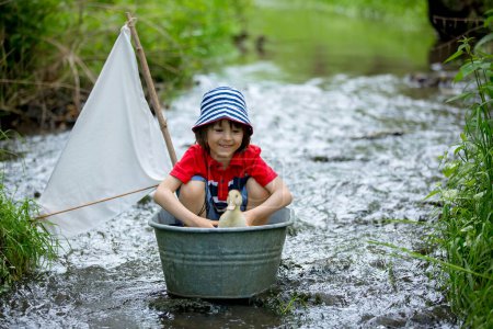Téléchargez les photos : Enfant mignon, garçon, jouer avec le bateau et les canards sur une petite rivière, voile et canotage. Enfant s'amuser, concept de bonheur d'enfance - en image libre de droit