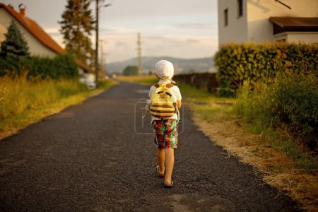 Foto de Beautiful child, boy with backpak walking on a little rural path on sunset, after rain, summertime - Imagen libre de derechos