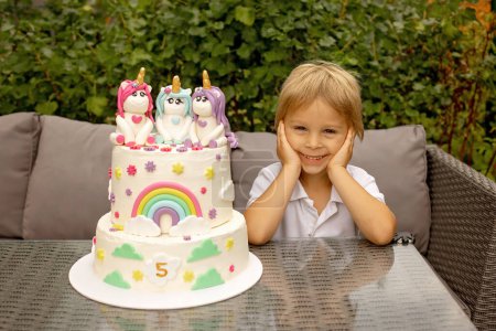 Foto de Cute five years old child, sitting in garden with big homemade cake with unicorns, celebrating birthday, beautifully decorated cake - Imagen libre de derechos