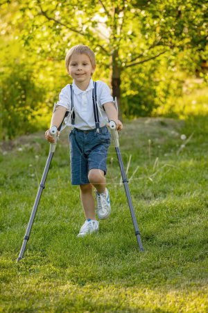 Foto de Lindo niño, niño, caminando con muletas en un jardín, con la pierna lesionada - Imagen libre de derechos