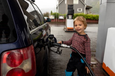 Téléchargez les photos : Sweet little child, blond boy, helping parents to put fuel in the car on a gas station - en image libre de droit