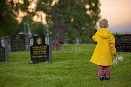 Foto de Sad little child, blond boy, standing in the rain on cemetery, sad person, mourning - Imagen libre de derechos