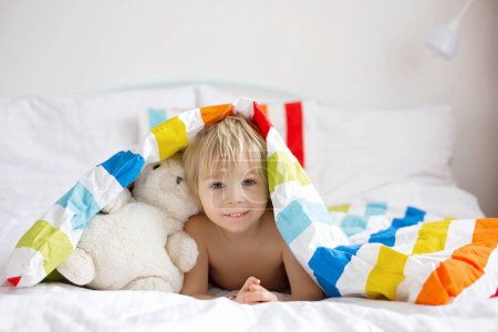 Téléchargez les photos : Bébé enfant heureux, garçon blond avec peignoir coloré, assis dans le lit avec jouet en peluche après la baignoire, souriant, literie colorée - en image libre de droit