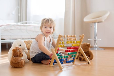 Téléchargez les photos : Enfant d'âge préscolaire blond, tout-petit, jouant avec un abaque à la maison, entouré d'ours en peluche - en image libre de droit