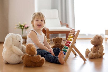 Foto de Dulce niño rubio preescolar, niño pequeño, jugando con ábaco en casa, rodeado de osos de peluche rellenos - Imagen libre de derechos
