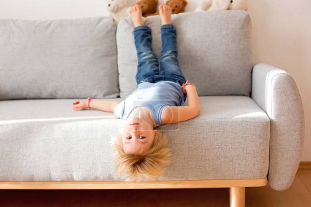 Téléchargez les photos : Enfant en bas âge, pendu à l'envers d'un canapé à la maison, souriant joyeusement - en image libre de droit