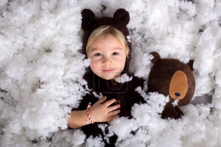 Photo for Cute blond toddler boy in knitted bear overall, holding teddy bear toy, isolated on gray background - Royalty Free Image