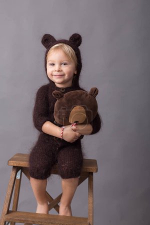 Téléchargez les photos : Mignon blond tout-petit garçon en tricot ours dans l'ensemble, tenant jouet ours en peluche, isolé sur fond gris - en image libre de droit