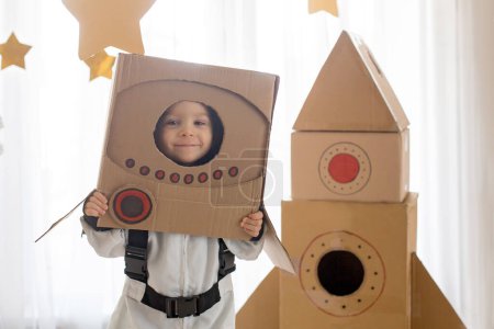 Téléchargez les photos : Petit garçon doux, habillé en astronaute, jouant à la maison avec une fusée en carton et un casque fait main de la boîte - en image libre de droit