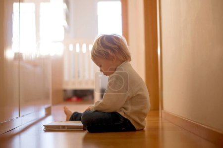 Foto de Hermoso niño rubio, acostado en el suelo en casa en el pasillo, libro de lectura, hermosa luz trasera - Imagen libre de derechos