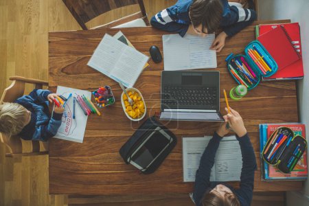 Téléchargez les photos : Trois enfants, assis autour de la table, écrivant des tâches scolaires pendant leurs études à domicile, tandis que l'école fermait à cause du coronavirus - en image libre de droit