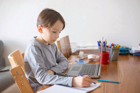Téléchargez les photos : Enfant à l'école, assis à la table avec ordinateur portable, écrivant des tâches scolaires pendant ses études à domicile, tandis que l'école fermait à cause du coronavirus - en image libre de droit