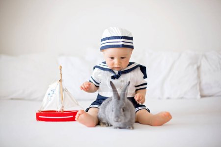 Téléchargez les photos : Bébé garçon mignon, vêtu de vêtements marins, jouant avec un bateau en bois et petit lapin mignon dans la chambre - en image libre de droit