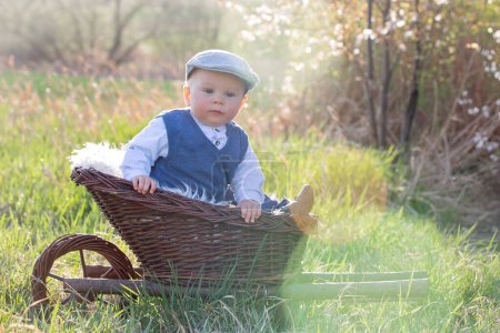 Photo for Cute little baby boy in trolley in a blooming garden with little rabbit and colorful easter eggs, smiling at camera - Royalty Free Image