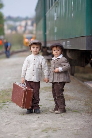 Foto de Dos chicos, vestidos con ropa vintage y sombrero, con maleta, en una estación de tren - Imagen libre de derechos