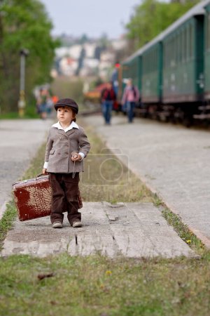Foto de Dos chicos, vestidos con ropa vintage y sombrero, con maleta, en una estación de tren - Imagen libre de derechos
