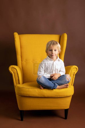 Téléchargez les photos : Enfant d'âge préscolaire en chemise blanche, vêtements décontractés, assis sur un fauteuil jaune sur un fond isolé, portrait élégant - en image libre de droit