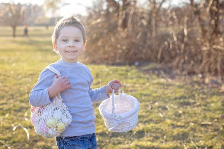 Téléchargez les photos : Petit garçon mignon, ramassant des œufs colorés de Pâques dans un panier dans le parc, printemps - en image libre de droit