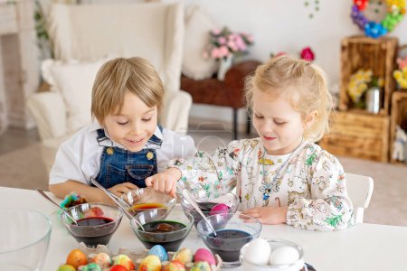 Foto de Feliz niñera, hermanos, huevos para colorear con aguas coloridas para Pascua en casa, sala soleada - Imagen libre de derechos