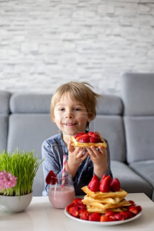 Photo for Sweet preschool child, boy, eating belgian waffle with strawberries and chocolate at home and driking smootie - Royalty Free Image