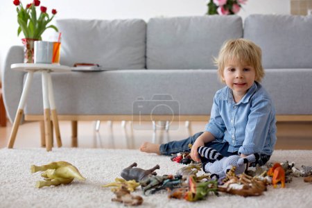 Téléchargez les photos : Enfant blond mignon, tout-petit garçon, jouant à la maison avec des dinosaures, assis sur le sol - en image libre de droit