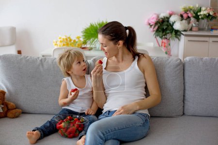 Téléchargez les photos : Mère et enfant, manger des fraises et s'amuser à la maison, assis sur le canapé - en image libre de droit
