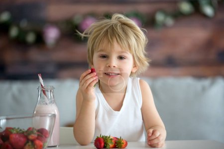 Photo for Cute toddler child, blond boy, drinking smoothie and easting strawberries at home - Royalty Free Image