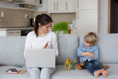 Foto de Madre, trabajando en su computadora portátil y tomando llamadas telefónicas, niño jugando junto a ella en casa mientras mamá tiene oficina en casa - Imagen libre de derechos