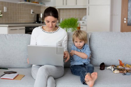 Photo for Mother, working on her laptop and taking phone calls, child playing next to her at home while mom having home office - Royalty Free Image