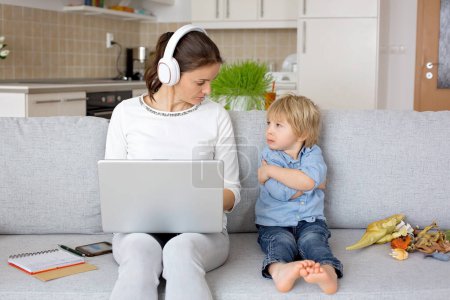 Téléchargez les photos : Mère, travaillant sur son ordinateur portable et prenant des appels téléphoniques, enfant jouant à côté d'elle à la maison tandis que maman ayant bureau à la maison - en image libre de droit