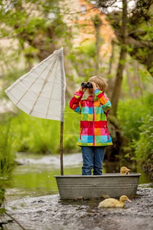 Téléchargez les photos : Enfant mignon, garçon en veste colorée, jouant avec bateau et canards sur une petite rivière, voile et canotage. Enfant s'amuser, concept de bonheur d'enfance - en image libre de droit