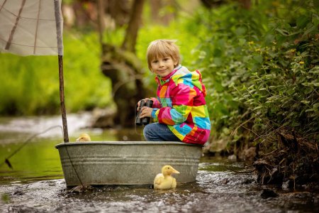 Foto de Lindo niño, niño con chaqueta de colores, jugando con el barco y patos en un pequeño río, vela y paseos en bote. Niño divertirse, concepto de felicidad de la infancia - Imagen libre de derechos