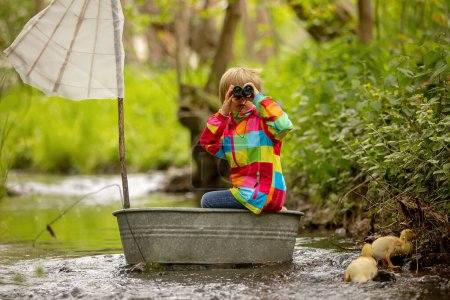 Téléchargez les photos : Enfant mignon, garçon en veste colorée, jouant avec bateau et canards sur une petite rivière, voile et canotage. Enfant s'amuser, concept de bonheur d'enfance - en image libre de droit