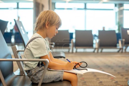 Téléchargez les photos : Enfant, garçon mignon, voyageant en famille, attendant à l'aéroport pour monter à bord de l'avion, dessin dans la salle d'attente - en image libre de droit
