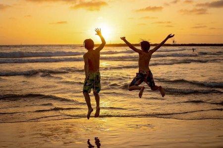 Téléchargez les photos : Heureux adolescents garçons, courir et jouer sur la plage au coucher du soleil, éclabousser l'eau et sauter sur le sable. Tel Aviv, Israël - en image libre de droit
