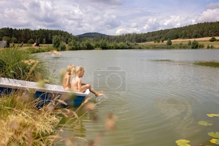 Téléchargez les photos : Beaux enfants blonds, garçon et fille, frères et sœurs, assis sur un bateau dans un lac, jouissant d'une atmosphère estivale incroyable ensemble - en image libre de droit