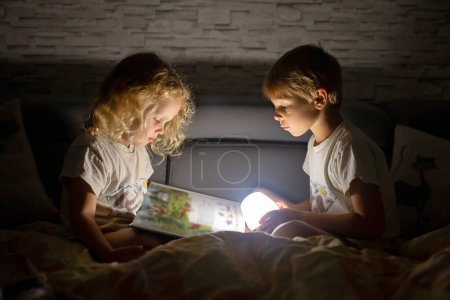 Foto de Dos niños lindos rubios, niño y niña, hermanos, acostados debajo de la cubierta en la cama, leyendo el libro junto con una pequeña luz, alegría y felicidad - Imagen libre de derechos