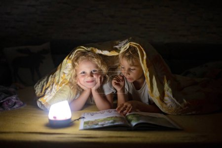 Téléchargez les photos : Deux blonds mignon enfant, garçon et fille, frères et sœurs, couché sous le couvercle dans le lit, livre de lecture avec une petite lumière, joie et bonheur - en image libre de droit