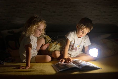 Photo for Two blond cute childrem, boy and girl, siblings, lying under the cover in bed, reading book together with small light, joy and happiness - Royalty Free Image