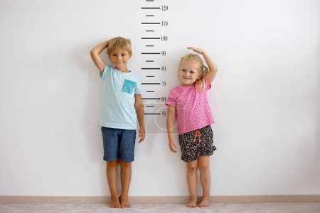 Photo for Little children, blond boy and girl, measuring height against wall in room, comparing themselves - Royalty Free Image